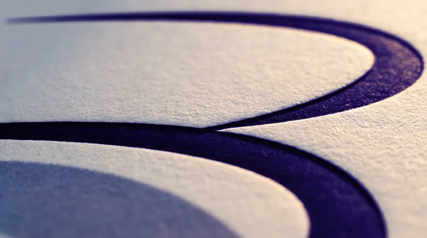 Gros plan d’un B calligraphié en bleu nuit, imprimé en relief sur un papier blanc texturé.